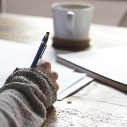 Photo showing a hand filling out paperwork with a coffee mug on the table