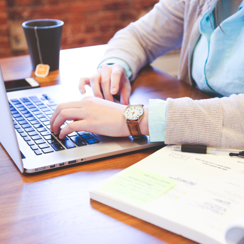 Tabletop showing student's hands typing on a laptop with book and cup of tea